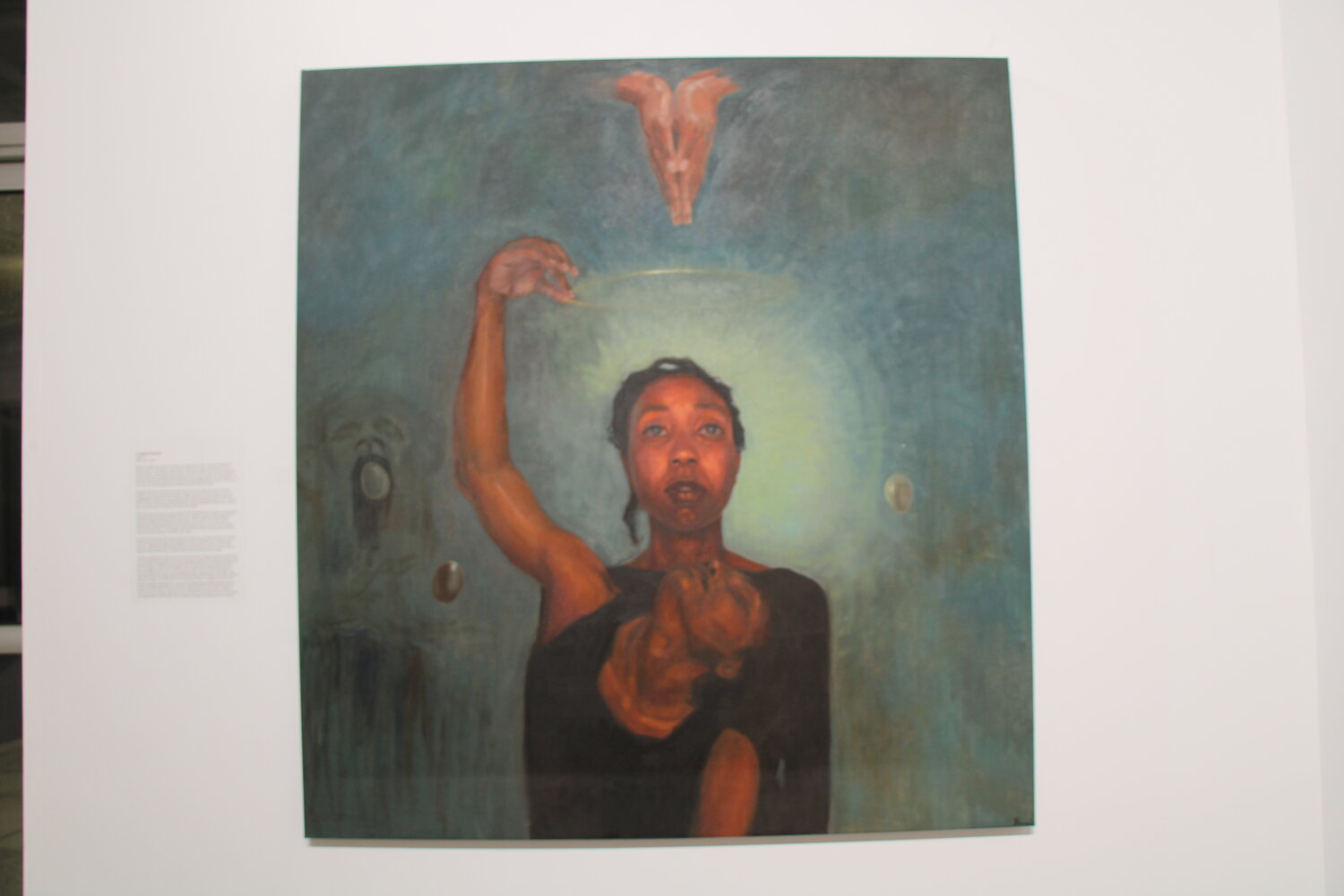 “Untitled” by Florence Wangui, a Kenyan artist has “largely gone about her work in quiet solitude.” The stark, yet warming portrait shows a young woman in flux with herself, possibly haunted with the vague figure on the left and hopefully absolved by the halo and praying hands above her.